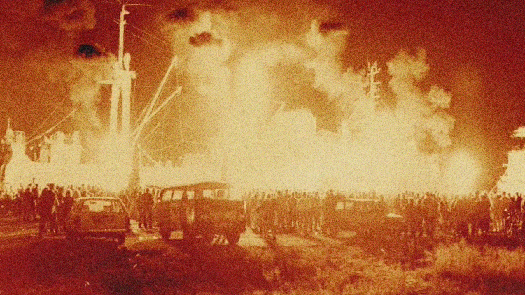 Eine Pyroshow vor der Stubnitz 1993. Die Silhouette des Schiffs ist vom Feuer erleuchtet. Rauchschwaden ziehen in den Himmel. Eine große Menge an Menschen schaut zu. Drei Autos stehen im Vordergrund.
