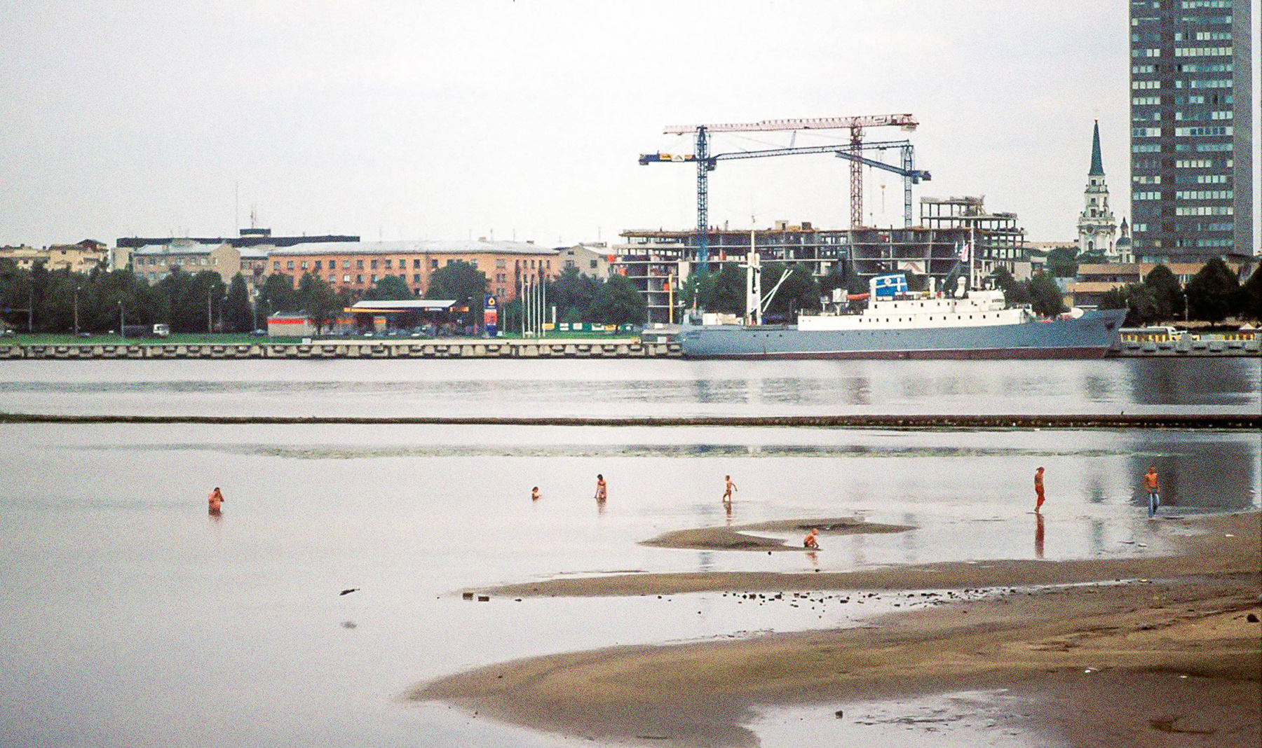 Die Stubnitz in Riga 2005. Im Vordergrund ein Stadtstrand mit badenden Menschen. Im Hintergrund Gebäude und Kräne.