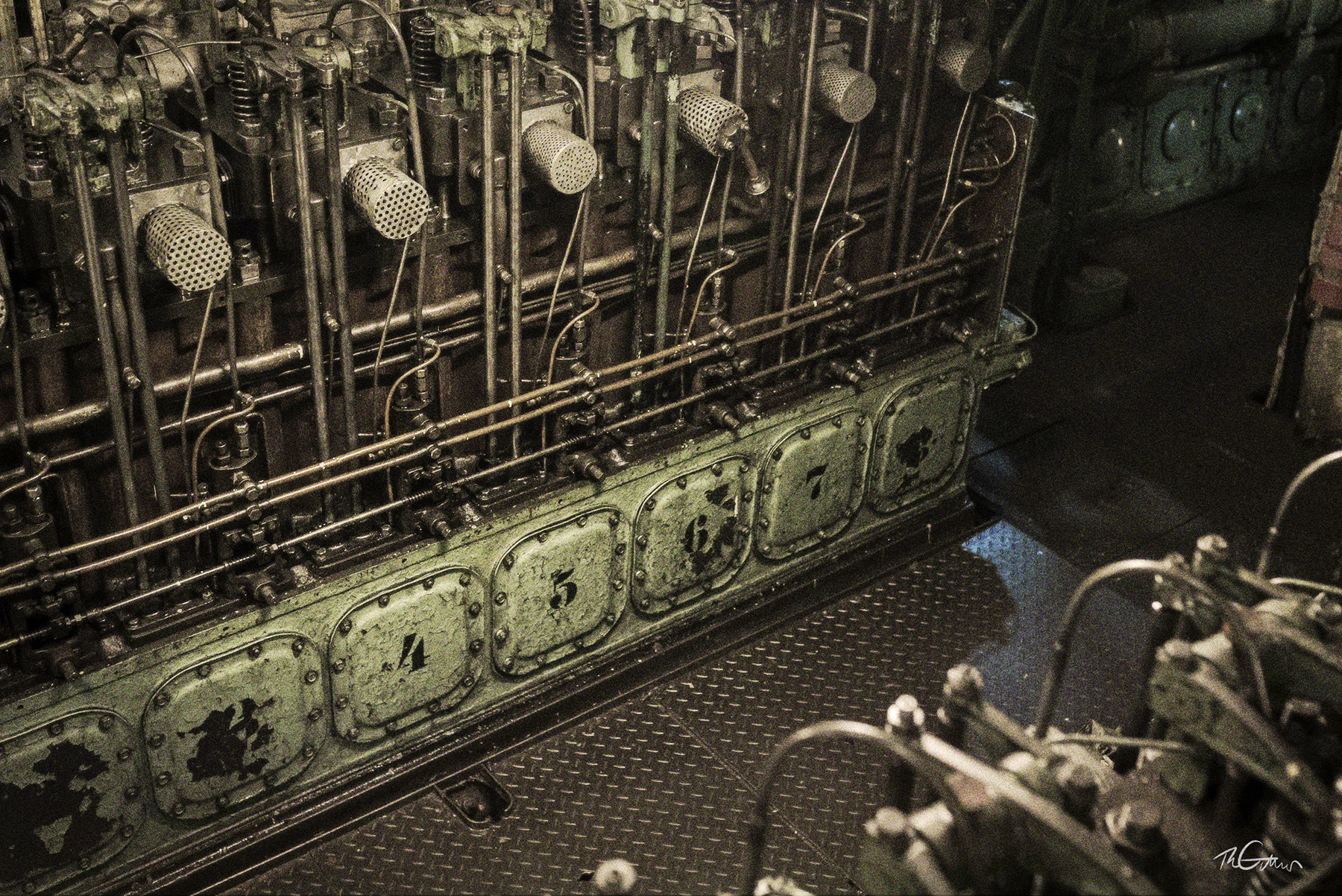 Die Hilfsmaschine im Maschinenraum. Es sind mehrere Zylinderköpfe und davor Bleche mit Nummern sichtbar.