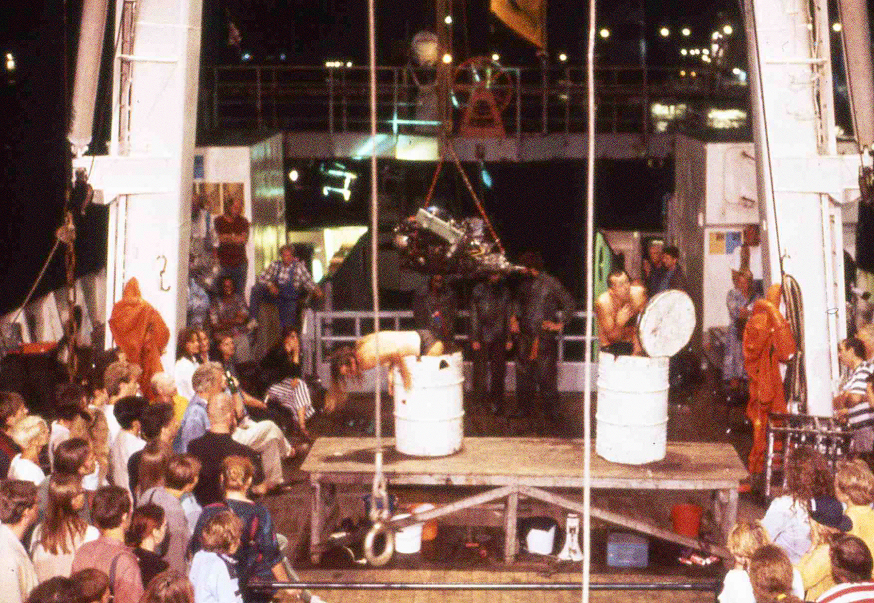 Eine Performance auf dem Achterdeck 1994 in St. Petersburg. Menschen klettern aus Metallfässern. Außenrum Zuschauer:innen.