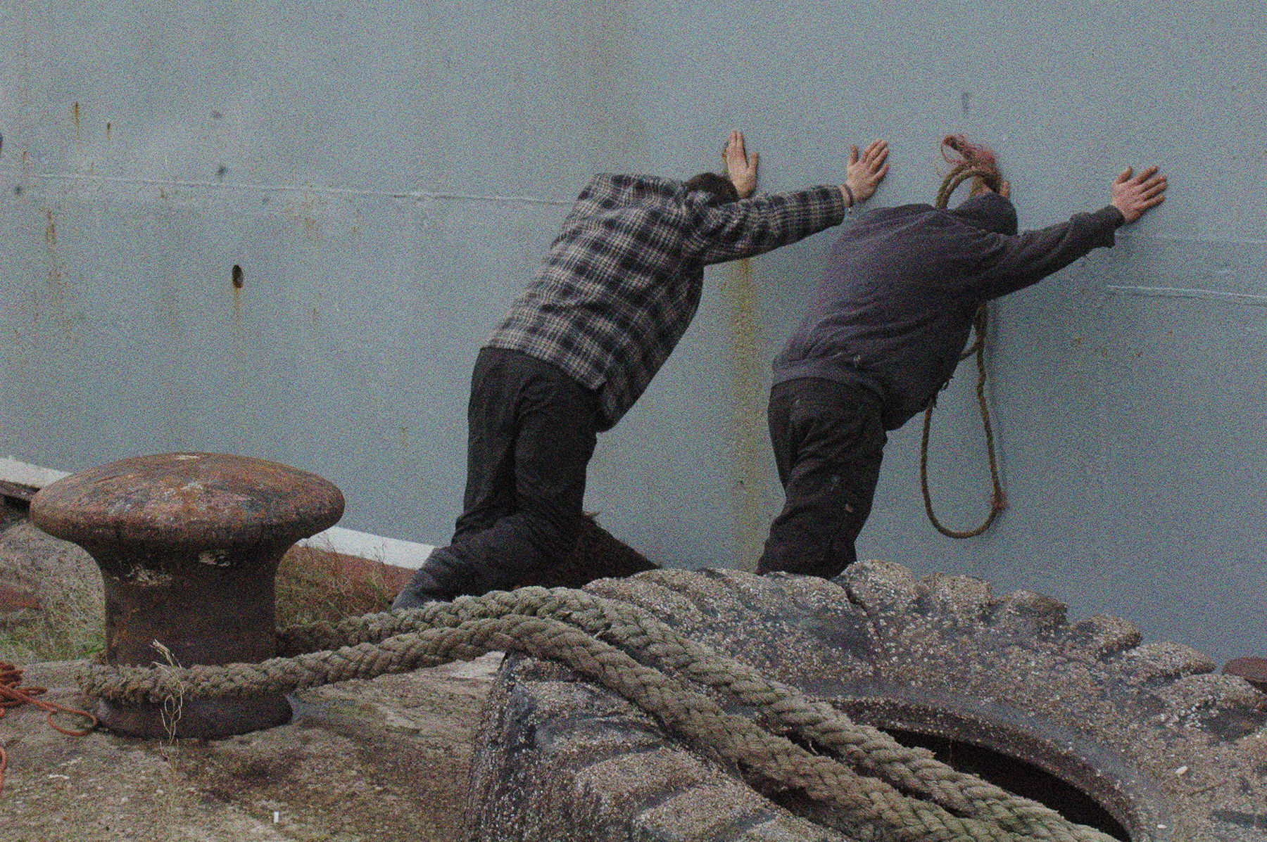 Die Stubnitz legt an. Nahaufnahme von zwei Menschen, die sich gegen die Schiffsseite stützen. Im Vordergrund ein großer Reifen und ein Poller mit einem Tau.