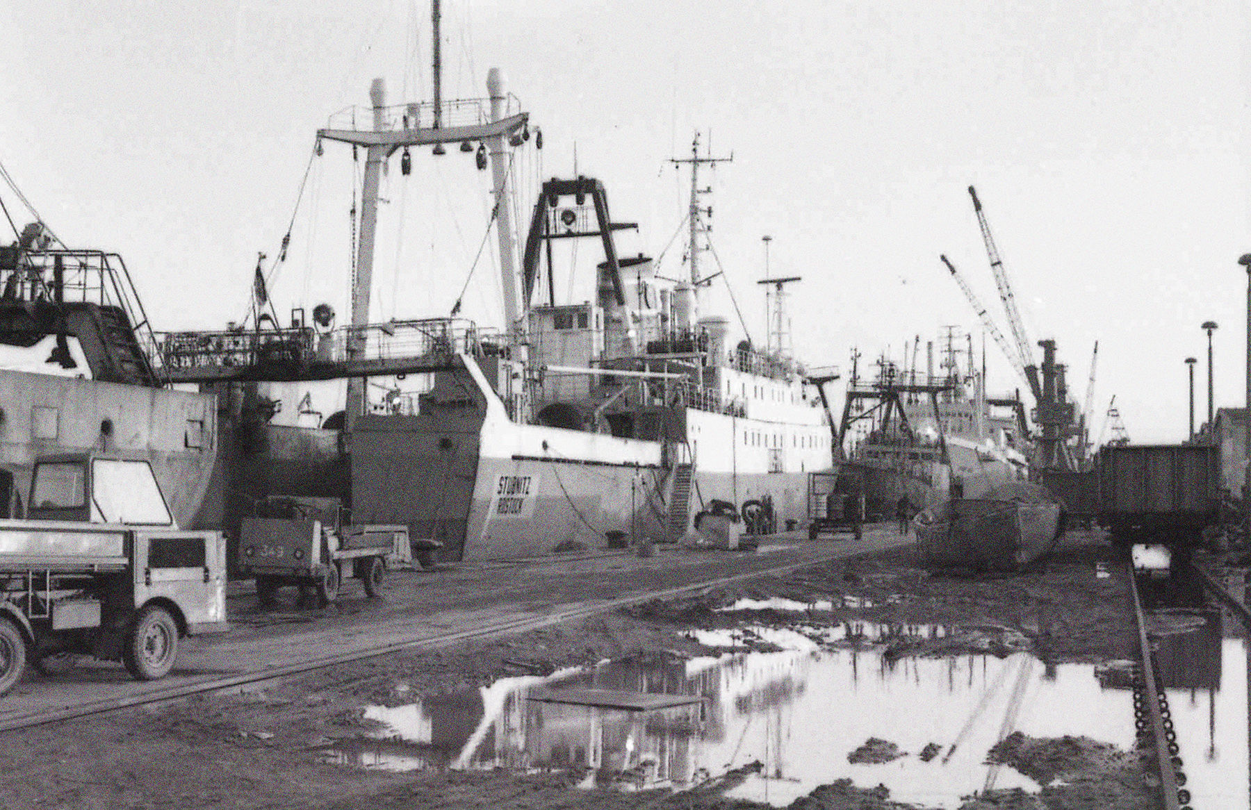 Die Stubnitz liegt im Rostocker Hafen. Vor und hinter dem Schiff liegen andere Schiffe an und Arbeitsfahrzeuge stehen an Land.