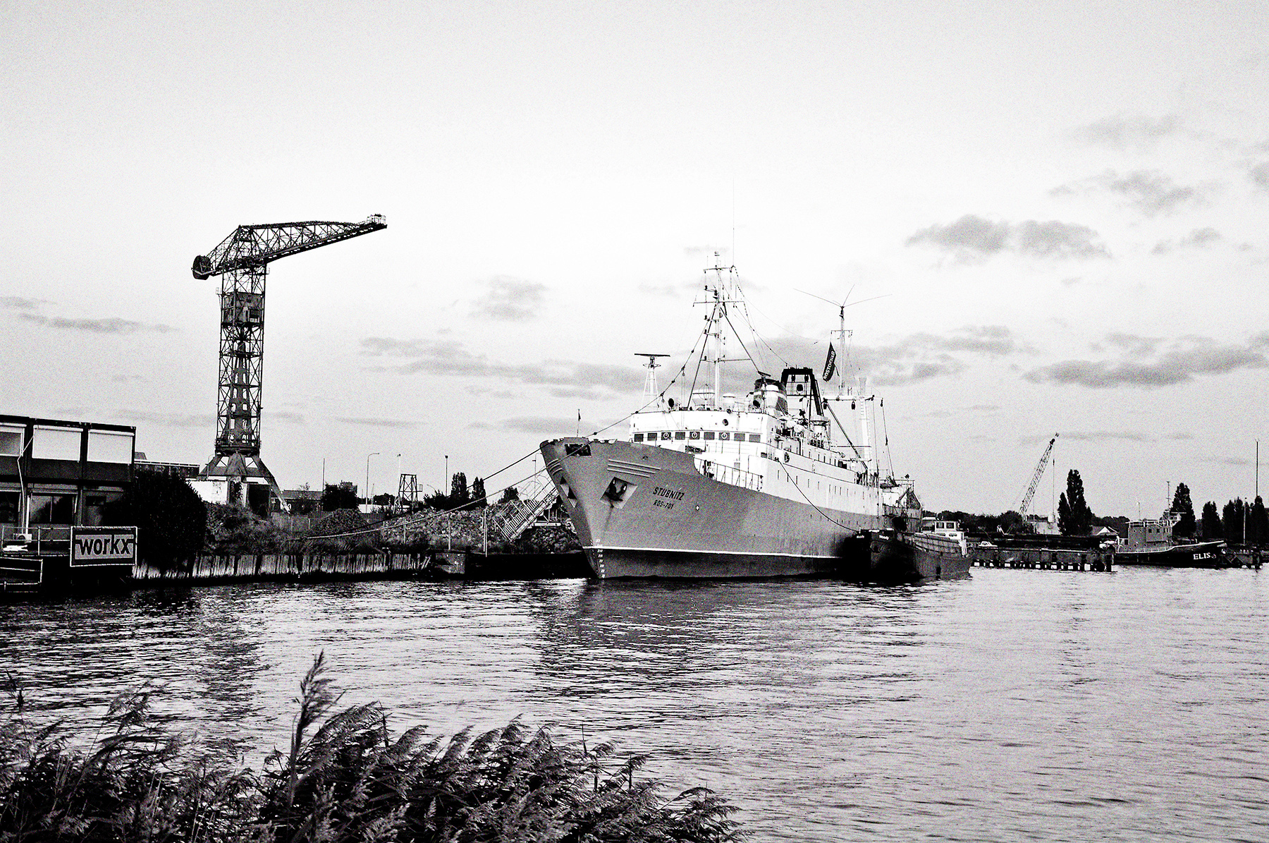 Die Stubnitz in Amsterdam 2009. Schwarzweiß Bild vom Ufer aus über das Wasser.Ikonografische Optik, Versorgungsboot liegt Backbord an, die Gangway geht an Land, ein großer Kran steht im Hintergrund.