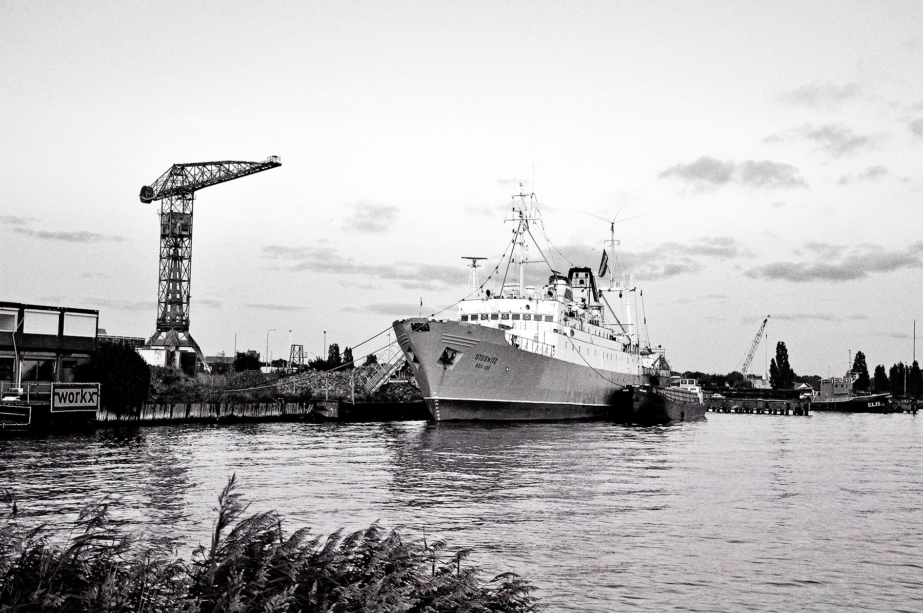 Die Stubnitz in Amsterdam 2009. Schwarzweiß Bild vom Ufer aus über das Wasser.Ikonografische Optik, Versorgungsboot liegt Backbord an, die Gangway geht an Land, ein großer Kran steht im Hintergrund.