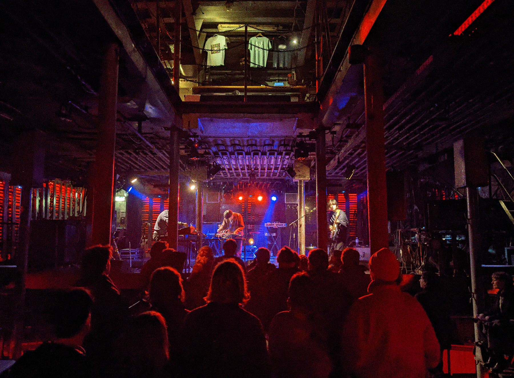 Ein Konzert von Maya Ongaku in Laderaum 4. Im Vordergrund steht im roten Licht das Publikum. Im Hintergrund steht die Band auf der Bühne in blauem Licht. 