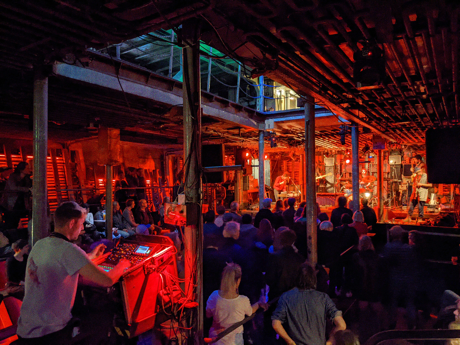 Konzert von Embryo in Laderaum 4. Die Band spielt auf der Bühne. Viele Menschen stehen vor der Bühne oder sitzen am Rand. Im Vordergrund sitzt eine Person am Mischpult.