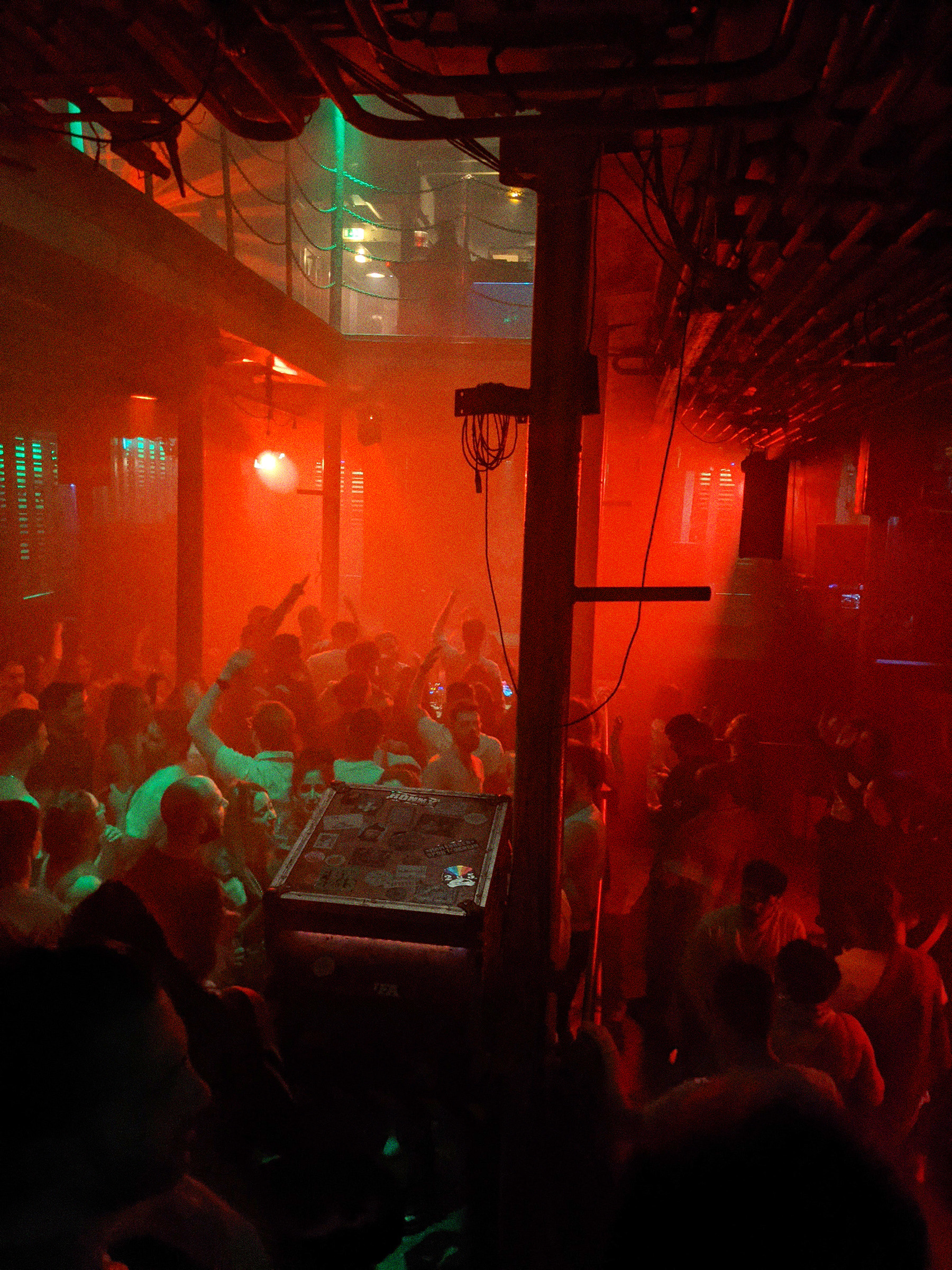 Eine Clubveranstaltung in Laderaum 4. Eine Menge tanzt in rotem Licht auf der Tanzfläche. Im Vordergrund das ungenutzte Konzert-Mischpult.