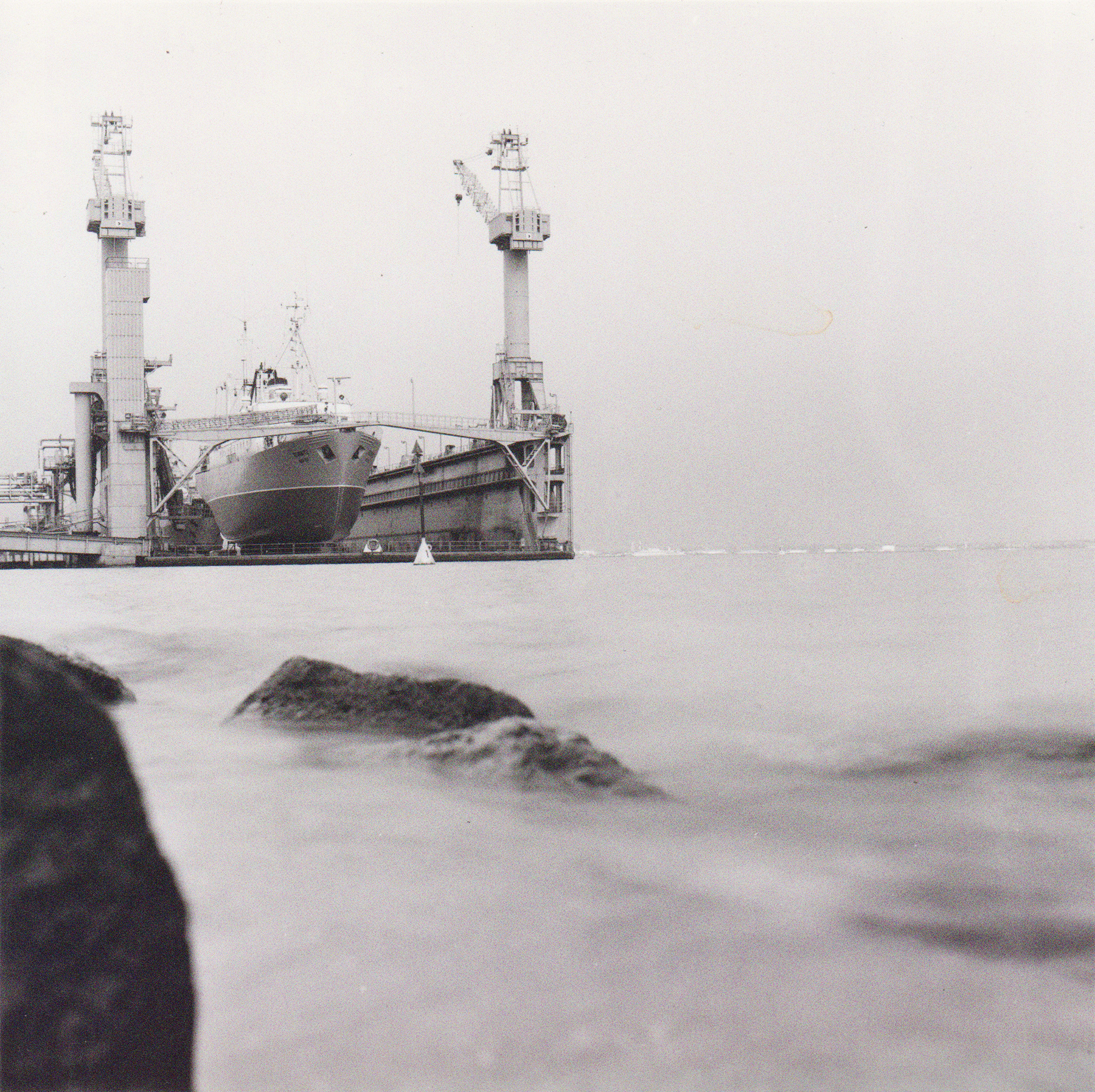 Die Stubnitz im Dock in der Warnowwerft in Rostock 1993. Werft und Schiff in der Ferne. Im Vordergrund Wasser und Steine.