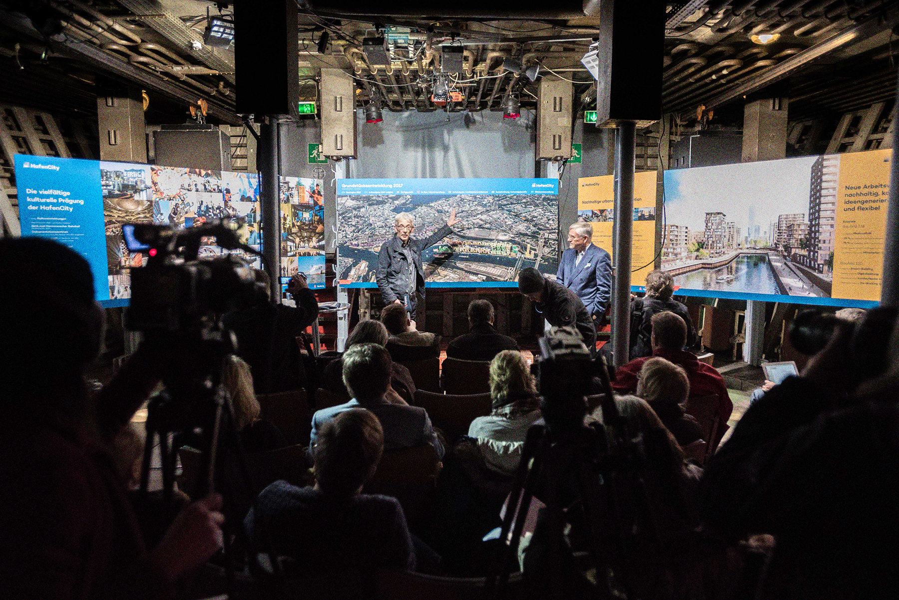 Eine Pressekonferenz in Laderaum 1. Im Vordergrund Menschen an Kameras von hinten, dahinter Stuhlreihen mit sitzenden Menschen. Ganz hinten im Bild zwei Menschen die vor Plakatwänden stehen und etwas erklären.