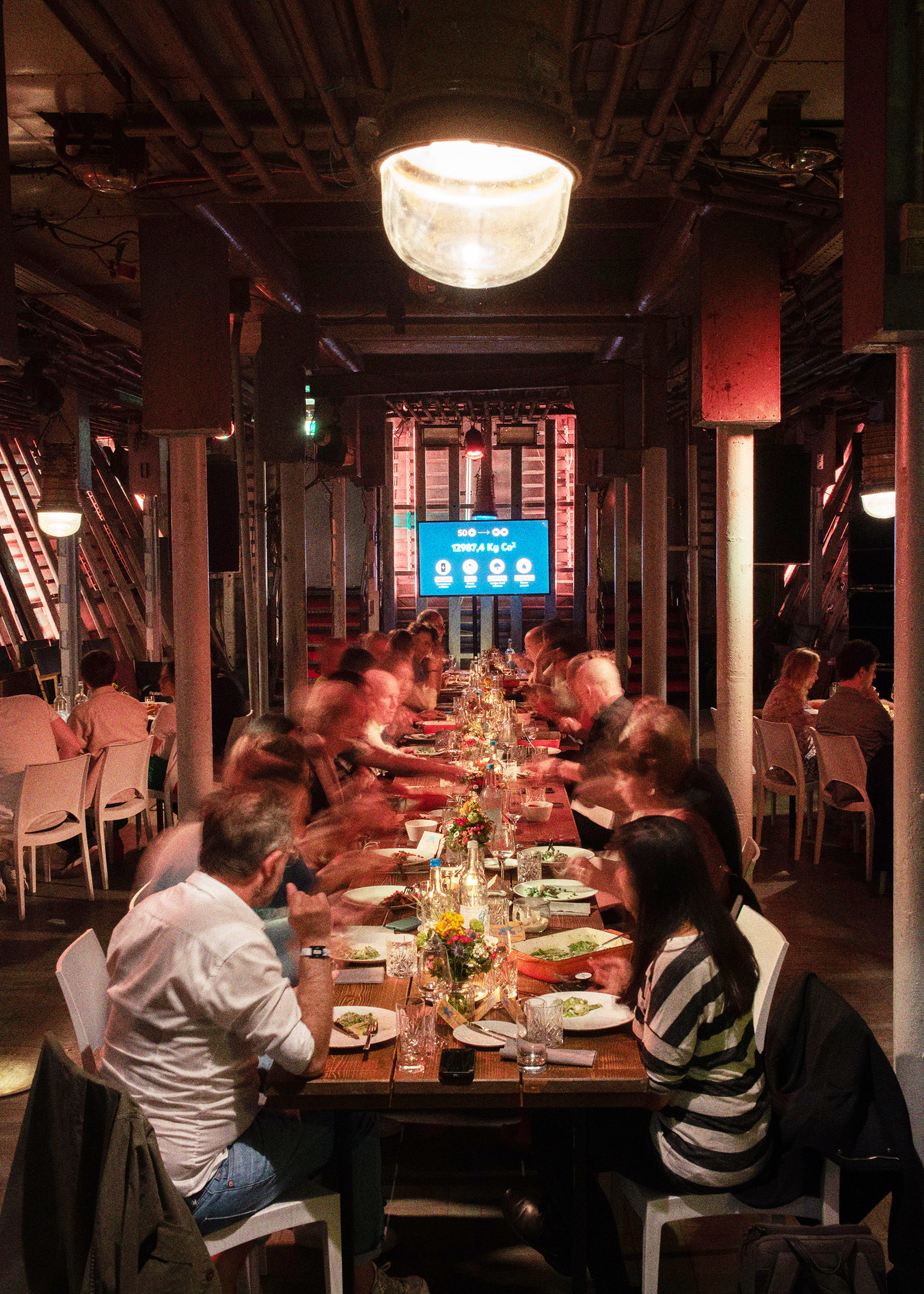 Eine lange, gedeckte Tafel in Laderaum 1. Menschen sitzen an der Tafel und essen. Am Ende der Tafel hängt ein Bildschirm.