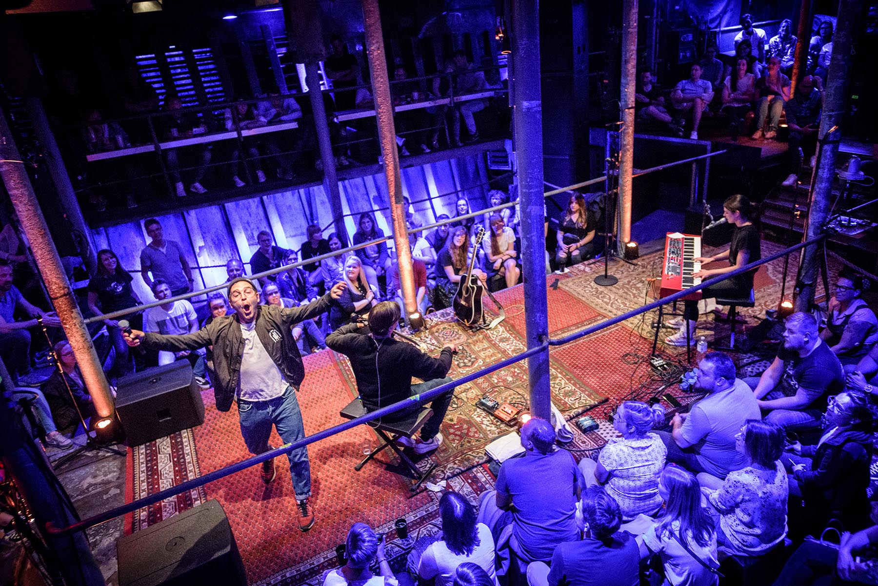 Blick von oben auf ein Konzert in Laderaum. Menschen sitzen und stehen auf allen Ebenen im Raum, er ist blau beleuchtet.